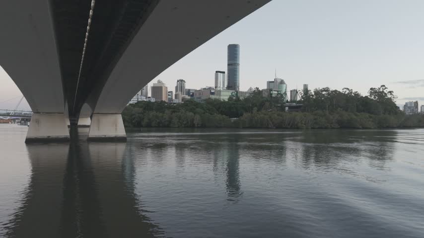 An aerial view passes beneath the concrete pylon of the Goodwill Bridge, the Brisbane skyline visible beyond at dawn.