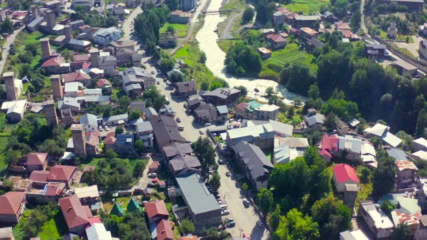 Mestia neighborhoods pass beneath forward aerial view beside a mountain river and historic tower houses, supporting city planning, river geography, heritage tourism and regional travel promotion.