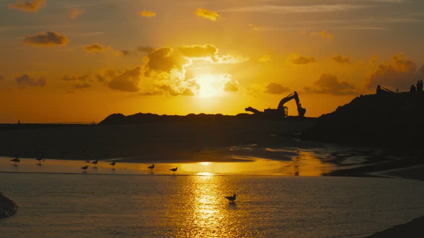 Sunset over beach with construction machinery silhouetted against the horizon, reflecting light on water and seagulls flying near the shore in a tranquil coastal scene