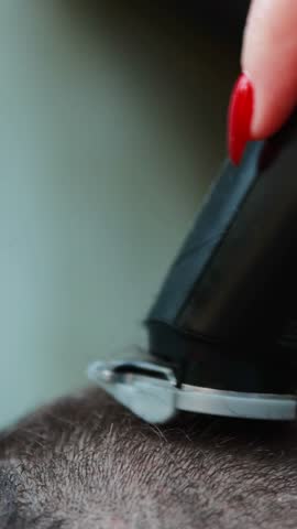 Close-up of a person with red manicure using a hair clipper to buzz-cut short brown hair. Hairdressing, grooming, and barber shop concept.