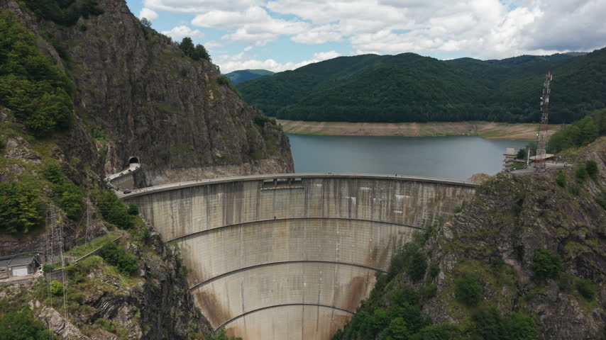Broad drone shot follows the arch wall toward Vidraru Lake, forest ridges and exposed reservoir shoreline.