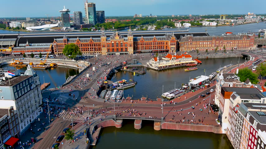 Aerial drone view of Amsterdam Centraal station with crowds of pedestrians and cyclists on Stationsplein, canal cruise boats on the Open Havenfront and the IJ river, Netherlands, in summer.