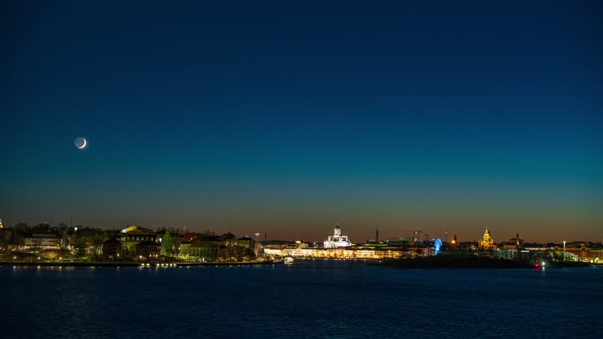View of the illuminated Helsinki skyline seen from Suomenlinna at night featuring a crescent moon over the waterfront in Helsinki, Uusimaa, Finland. Timelapse.