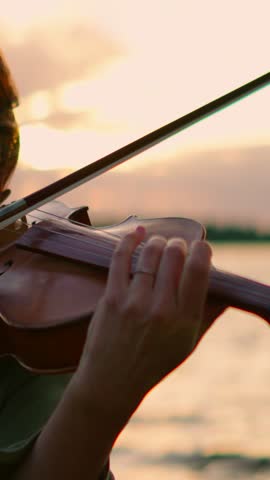 Close-up of a violinist playing soulful music by the water at sunset, focusing on the delicate hand positions and emotional expression of the performance