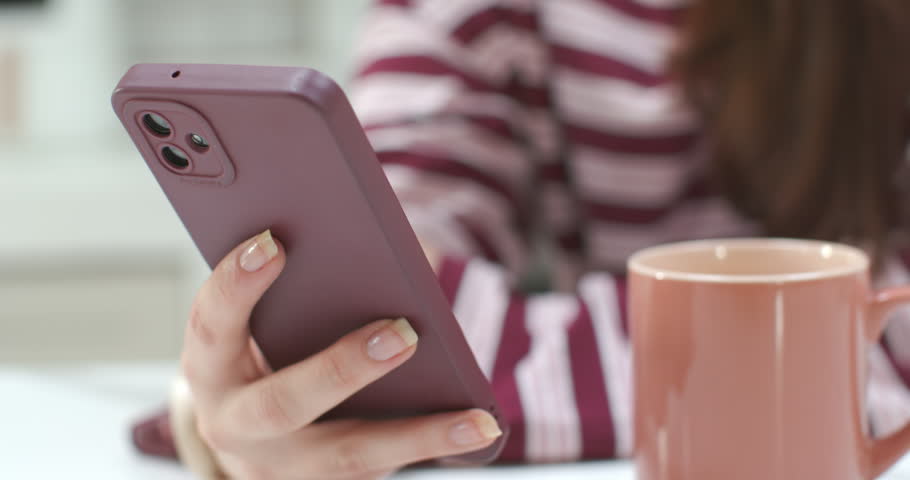 Woman holding purple phone by mug in kitchen, camera pulling back revealing her smiling at message. Smartphone, delight, home, countertop, stripedtop, nosepiercing, headband