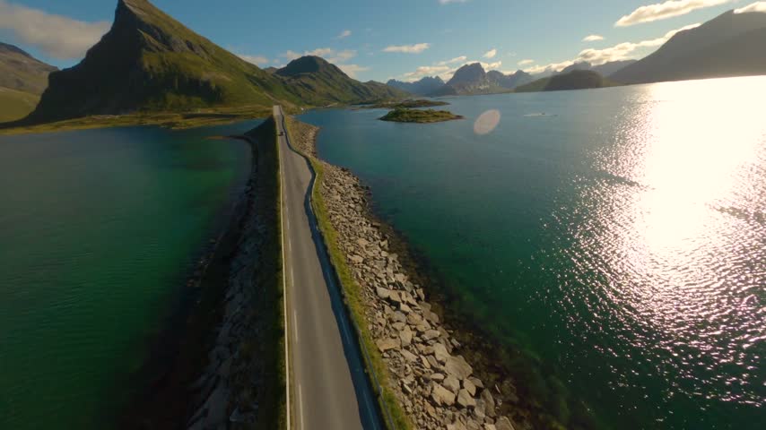 FPV aerial view of the scenic road on a causeway surrounded by turquoise water and dramatic mountains in Lofoten, Nordland, Norway.