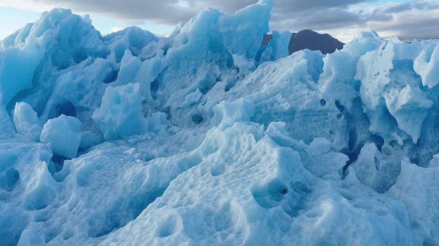 Large blue-white iceberg drifts across Jokulsarlon Glacier Lagoon in southeast Iceland, with sculpted ice formations and distant mountain ridges beyond the glacial water, pullback.