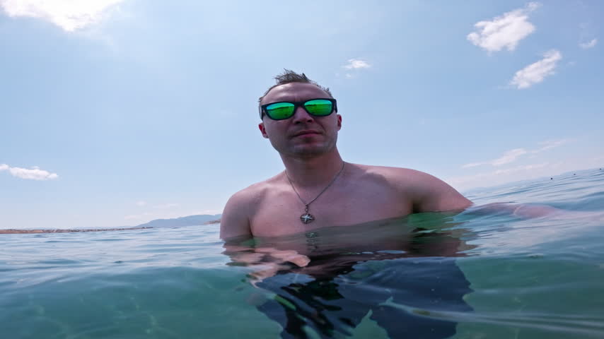 Bare-chested man in sunglasses swims underwater with an action camera in clear turquoise Greek sea, creating playful holiday POV.