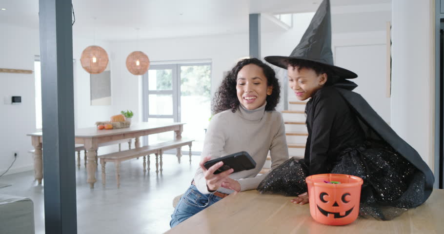 African American mom holding phone, child in witch hat leaning, taking pics at counter with pumpkin. Selfie, family, costume, halloween, kitchen, daylight, affection