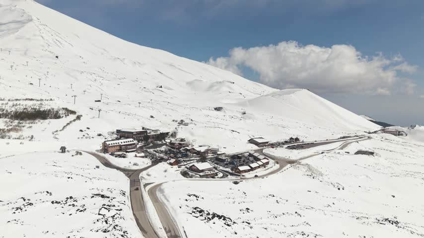 High-angle drone shot of the visitor center, restaurants, and parking area at the foot of Mount Etna craters in spring snow.