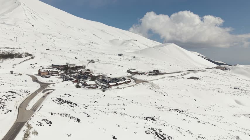 High-angle drone shot of the visitor center, restaurants, and parking area at the foot of Mount Etna craters in spring snow.