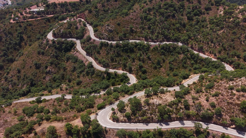 Winding mountain road with sharp hairpin bends cutting through dense green Mediterranean hillside vegetation viewed from above in summer
