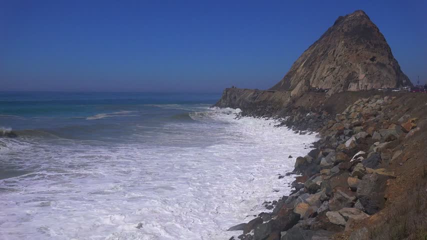 Large ocean waves crashing against rocky shoreline in Morro Bay California with scenic mountain backdrop in slow motion cinematic clip.