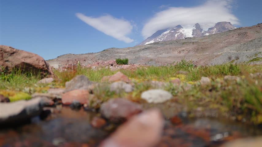 Low angle view across mossy rocks and a shallow alpine stream toward the glaciated summit of Mount Rainier National Park Washington crowned by a lenticular cloud under deep blue summer sky.