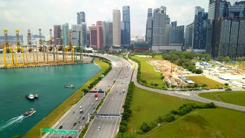 Aerial view Singapore cityscape showcases skyscrapers with bustling container port and cranes, busy highway traffic. Boats navigate turquoise waters.