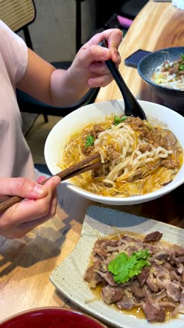 Close-up of hands using chopsticks and spoon to eat traditional Taiwanese noodles in Taiwan.