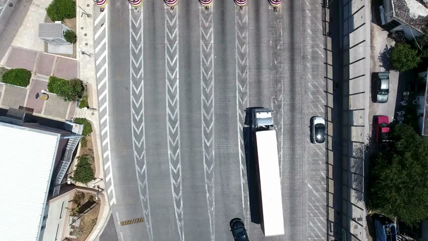 High angle drone view shows diverse traffic including cars and a large semi truck moving through a modern multi-lane highway toll plaza with numerous payment booths.