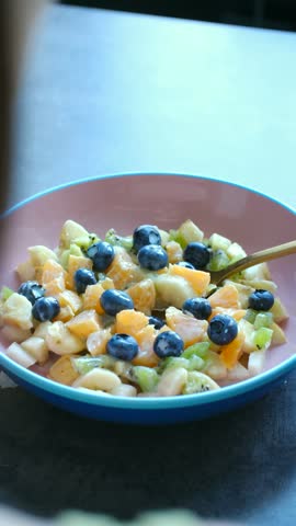 Person mixing fresh vibrant fruit salad with spoon in pink bowl, healthy meal full of blueberries, kiwi, banana, orange for delicious breakfast or dessert. Healthy fruit salad breakfast bowl mixing