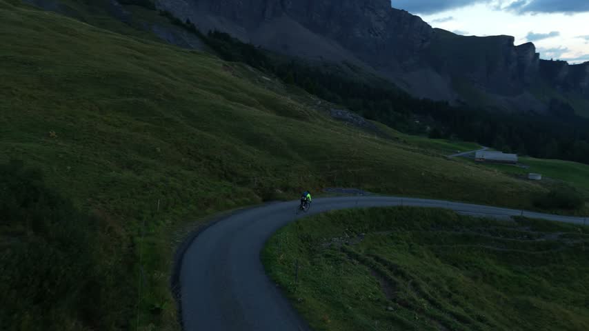 Drone follows a road cyclist descending a steep mountain road before sunrise, with alpine peaks and distant villages visible across the valley in low morning light.
