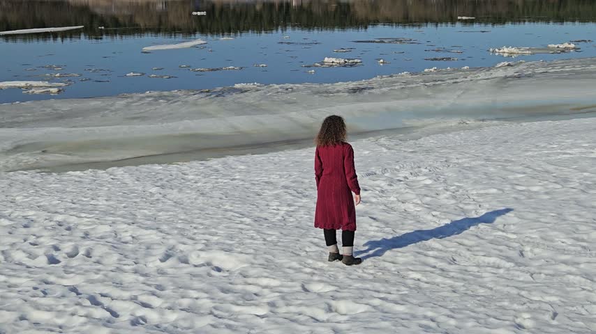Child in red coat stands on snowy beach beside icy river water. Footprints mark the snow near the frozen shoreline. Reflections ripple across the partially ice-covered surface