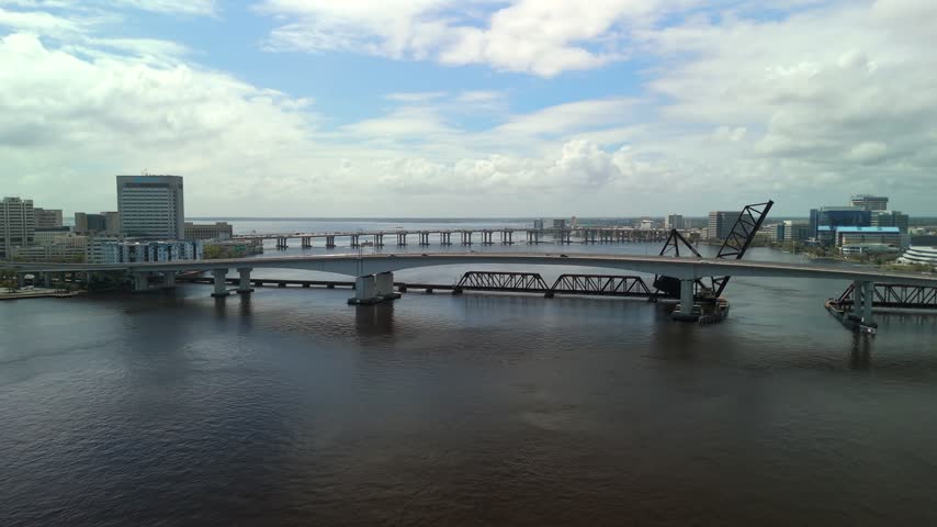 Drone view of Jacksonville, Florida, showing multiple bridges across the St. Johns River, waterfront buildings, and the urban skyline beneath dramatic clouds