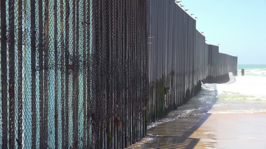 Waves crash against the border wall separating the United States and Mexico, a powerful symbol of immigration and national security.