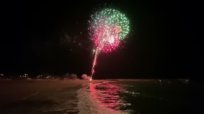 Colorful firework show on night beach with ocean waves reflection and cinematic smoke trails.