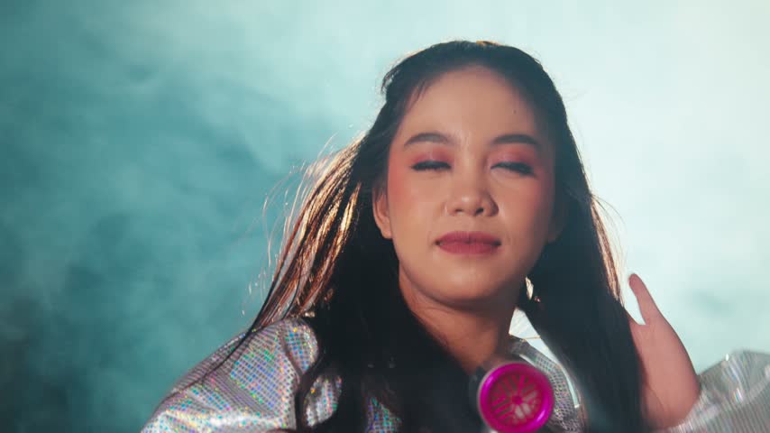 A young woman poses confidently while using a pink hair dryer to blow her hair, surrounded by thick white smoke and dramatic studio lighting.