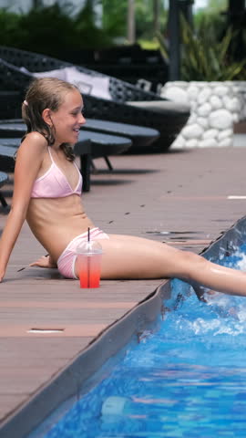 Vertical video. Teenage girl in a pink two-piece swimsuit sitting on the pool edge swinging her legs in the cool water, with a strawberry cocktail with ice placed next to her on the deck