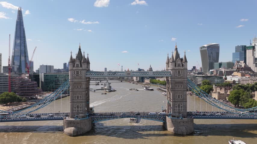 Aerial view captures Tower Bridge crossing the River Thames in London on a clear sunny afternoon while boats travel below and traffic moves across the bridge near famous tall city buildings