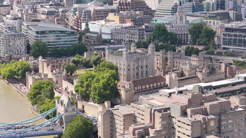 Tower of London and historic Tower Bridge stand near the River Thames during a bright sunny afternoon in England as traffic flows across the bridge and people walk through the grounds below