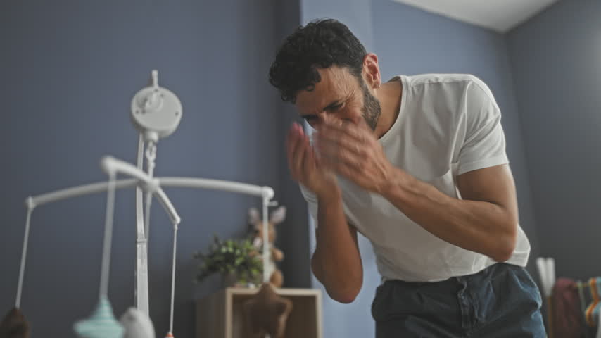 Man covers face with hands over crib and hanging mobile in a nursery building, leaning and peeking between toys; fatigue bonding care.
