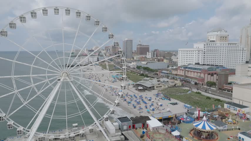 Steel Pier Ferris wheel towers over Atlantic City beachfront amusement attractions, aerial medium reveal of umbrellas behind promenade