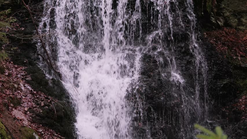 Multi-tier woodland waterfall cascading over mossy dark rock face in autumn forest, with silky white water, wet stone textures, fallen leaves, and calm natural scenery