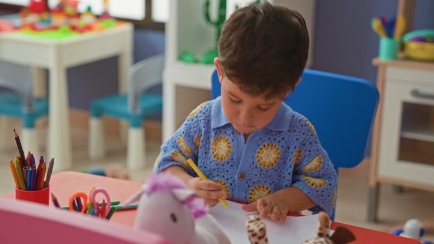Boy cutting paper with orange safety scissors, hand positioned on paper at a preschool table with crayons and toys visible; concentration.