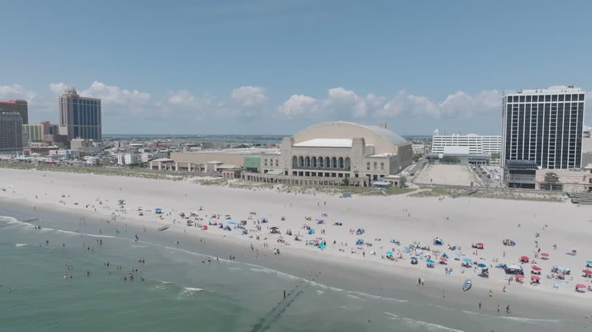 Boardwalk Hall fronts busy Atlantic City beach beside hotels and Atlantic Ocean, aerial panoramic orbit above people playing in water