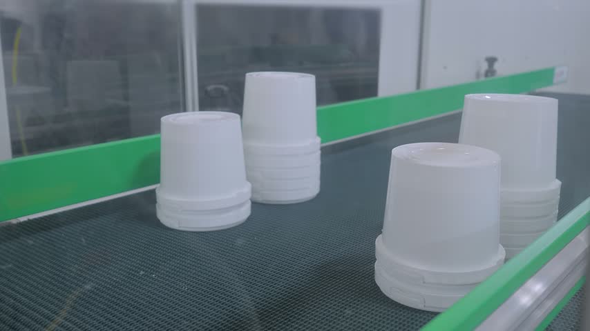 Stacks of white plastic yogurt containers are moving on conveyor belt of plastic injection molding machine with pick and place robotic arm manipulator at exhibition, factory - close up view