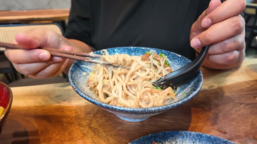 Close-up of hands using chopsticks to lift Ma Jiang Mian noodles from a bowl.