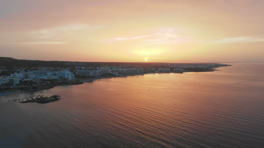 Aerial view of sunset over Ayia Napa coastal town in Cyprus featuring calm sea waters and orange sky cinematic drone footage.