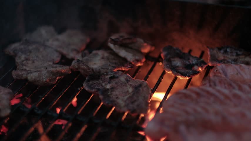 Various cuts of marinated meat and sausages cooking on a dark metal grill over glowing hot coals