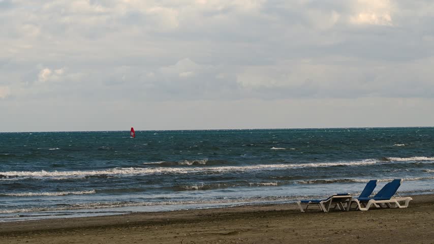 Windsurfer gliding on wavy sea near empty sandy beach with sun loungers on windy day.