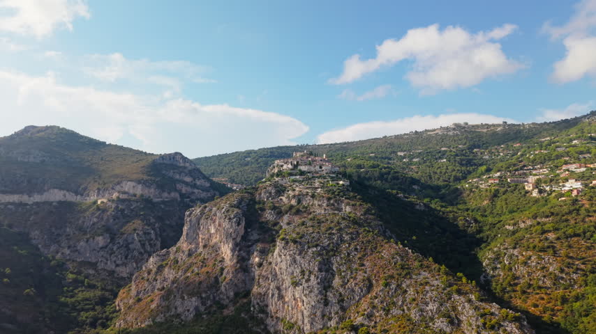 Eze Village medieval town perched on mountaintop, aerial wide shot over French Riviera valley with forested hills and cliffs, France scenic landscape.
