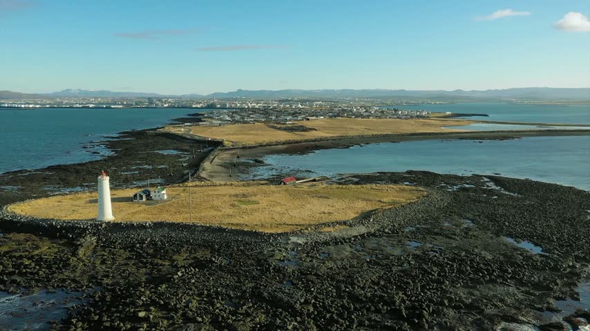 Drone video of Grotta Lighthouse on a small island near Reykjavik, Iceland, showing the coastal landscape, ocean, rocky shore, lighthouse tower, and clear sunny weather.