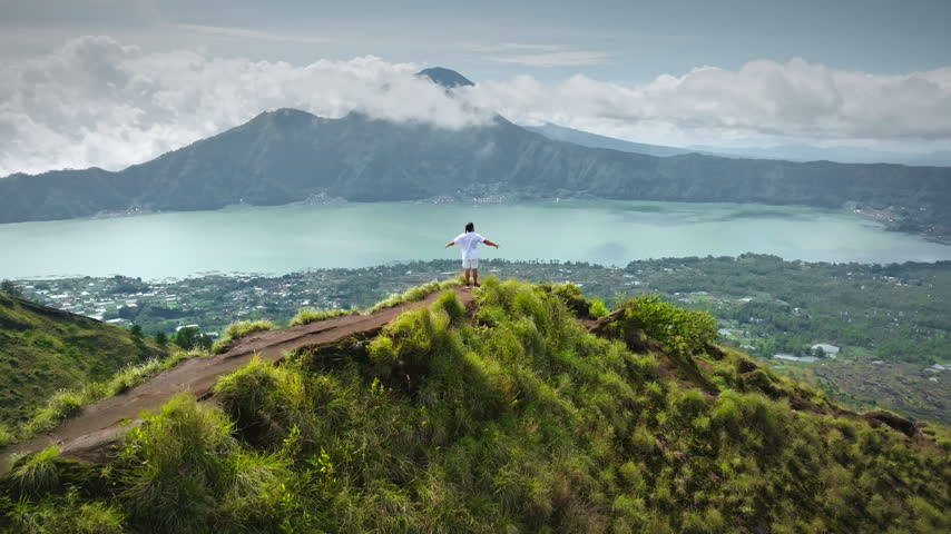 Person on a mountain peak raising arms in triumph and enjoying expansive panoramic view of lake Batur and surrounding landscape, symbolizing freedom, achievement, and travel adventure on Bali island