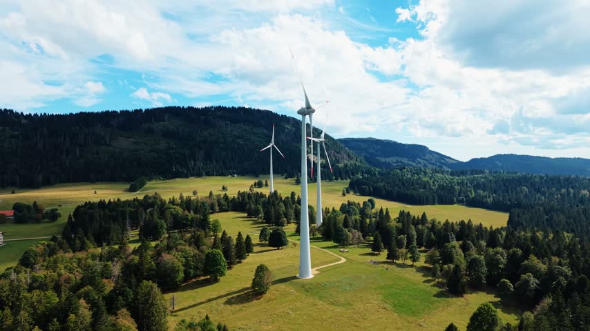 Aerial view of Sainte-Croix wind farm with large turbines set amidst lush green fields and dense pine forests under a bright sky in Sainte-Croix, Vaud, Switzerland.