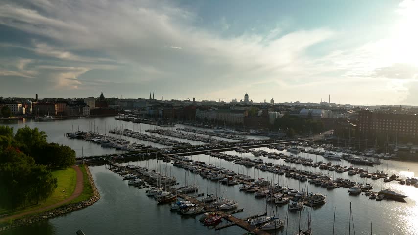 Drone panorama over a marina near Helsinki with numerous yachts and small boats at the docks and historic city houses visible beyond the calm coastal water