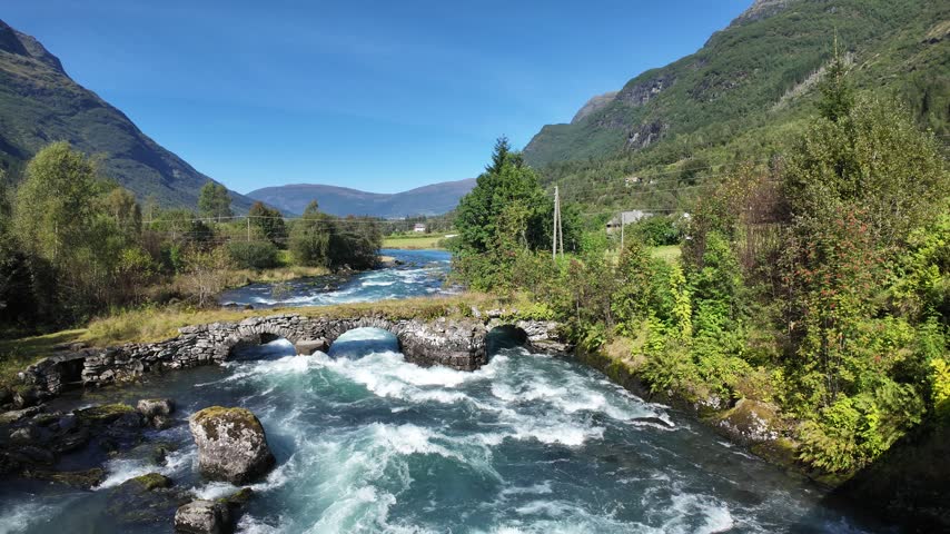 Looking over fast flowing water moving under a historic arched bridge. The bridge has grass on top, there is lush green foliage all around and dramatic mountain scenery in the background. The water is flowing towards Lake Floen in rural Norway.