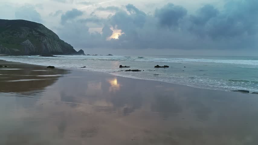 Aerial view of the Atlantic Ocean coastline with wet sand reflecting the cloudy sky and sunset light along the cliffs of Portugal.