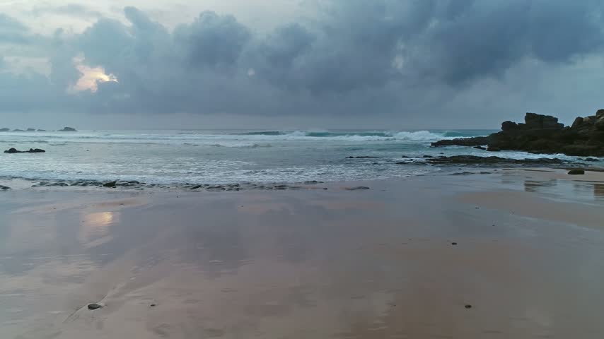 Aerial view of Atlantic Ocean waves crashing on the sandy beach with rocky formations under a dramatic cloudy sky in Portugal.