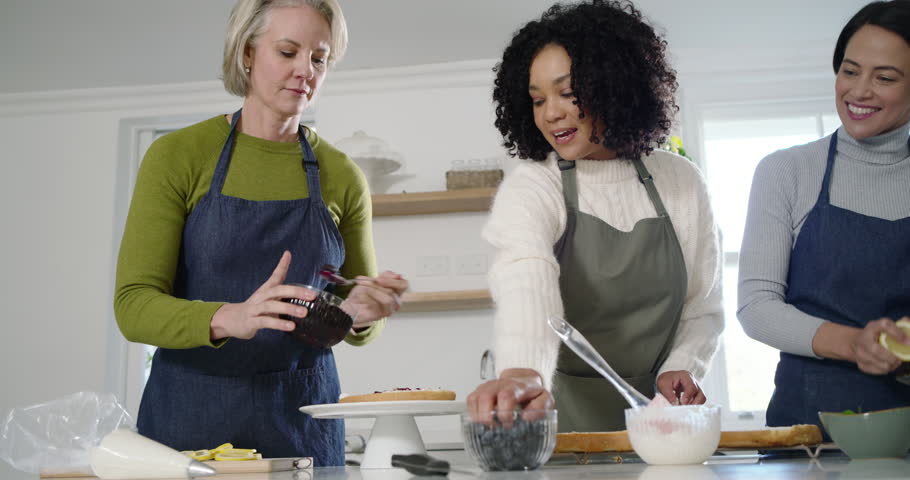 Diverse female friends with bowls assembling layer cake at kitchen island, frosting, zesting lemon. Baking, dessert, decorating, blueberries, plating, teamwork, collaboration